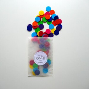 Może przedstawiać: Biała papierowa torebka wypełniona kolorowymi k&oacute;łkami konfetti z filcu. Torebka ma etykietę z napisem "Konfetti" i małym symbolem serca.