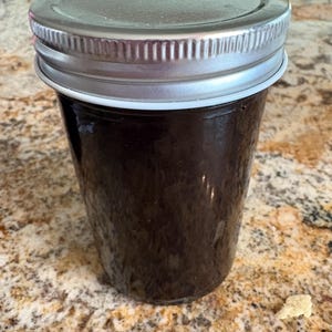 Peut inclure: Un pot en verre transparent avec un couvercle argenté, rempli d'une substance brun foncé. Le pot est posé sur un comptoir moucheté brun clair. Le couvercle est scellé, suggérant que le contenu est conservé.