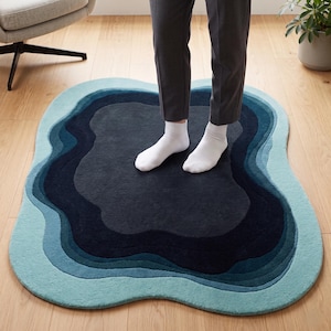 May include: An irregular, organic-shaped area rug featuring concentric, wavy layers of colour. The design transitions from a dark charcoal centre through shades of deep navy, teal, and light blue, creating a layered, topographical appearance.