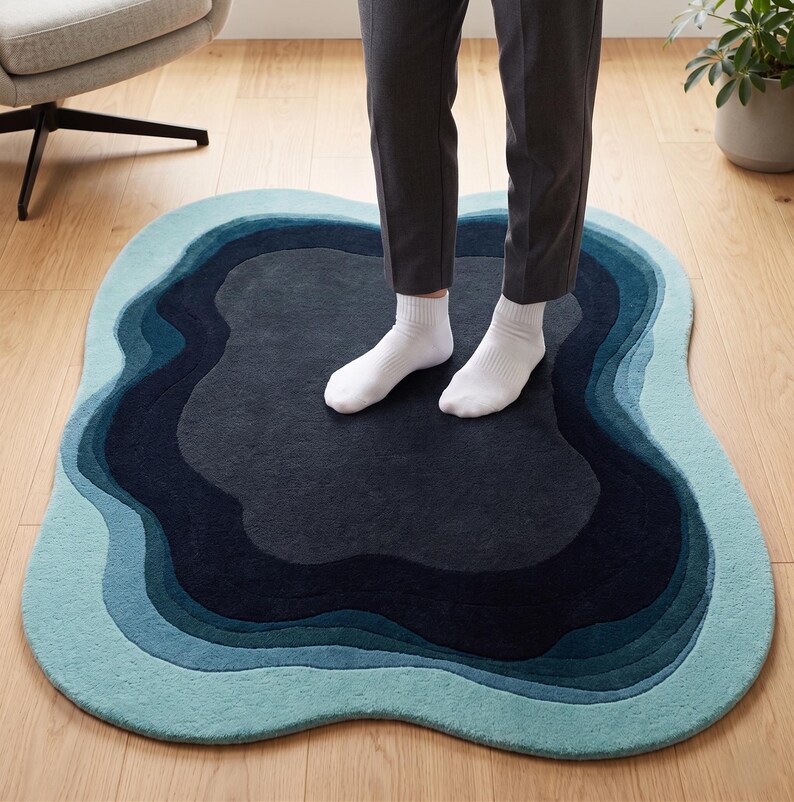 May include: An irregular, organic-shaped area rug featuring concentric, wavy layers of colour. The design transitions from a dark charcoal centre through shades of deep navy, teal, and light blue, creating a layered, topographical appearance.