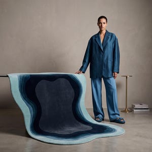 May include: A large, irregularly shaped area rug featuring concentric, organic layers of blue, ranging from light sky blue at the edges to deep navy in the centre. The rug has a soft, plush texture and a smooth, flowing, wave-like design.