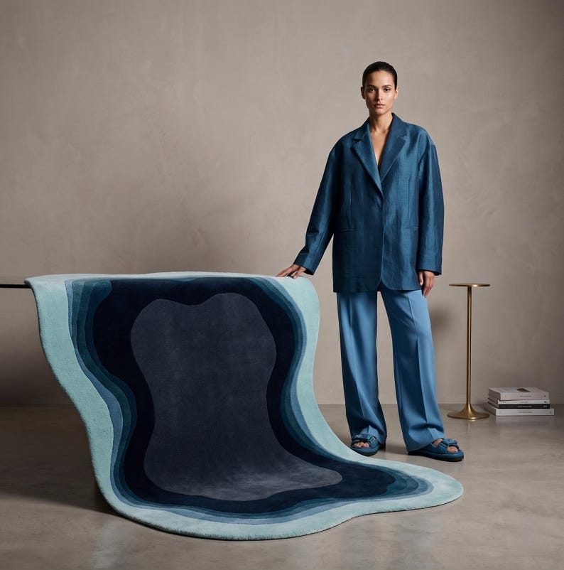 May include: A large, irregularly shaped area rug featuring concentric, organic layers of blue, ranging from light sky blue at the edges to deep navy in the centre. The rug has a soft, plush texture and a smooth, flowing, wave-like design.