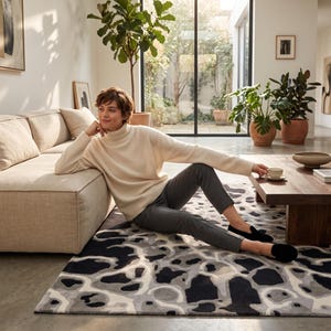 Puede incluir: Un su&eacute;ter de cuello alto color crema de manga larga, combinado con pantalones gris oscuro y zapatos negros sin cordones. El conjunto se lleva puesto mientras se est&aacute; sentado sobre una alfombra rectangular con un patr&oacute;n abstracto de formas negras, grises y blancas.