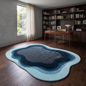 May include: An oval-shaped area rug featuring concentric, wavy layers of colour in shades of light blue, teal, navy, and dark grey. The rug has a soft, textured surface and is placed on a dark wood floor in a room with a desk and bookshelf.