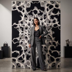Puede incluir: Un blazer gris carb&oacute;n y pantalones anchos a juego sobre una camiseta negra. El conjunto se sit&uacute;a frente a un gran tapiz de pared rectangular con un patr&oacute;n org&aacute;nico abstracto en tonos negro, gris y blanco.