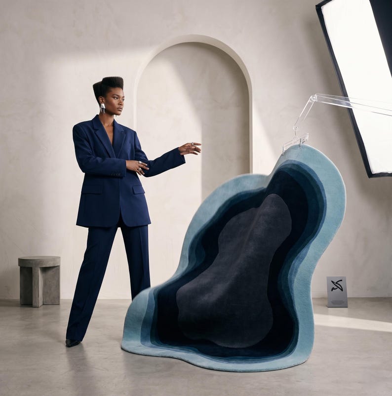 May include: A navy blue blazer and matching trousers paired with an irregular, organic-shaped rug featuring concentric layers of blue and teal gradients. The rug displays a deep central dark blue core transitioning outward to lighter shades of blue.