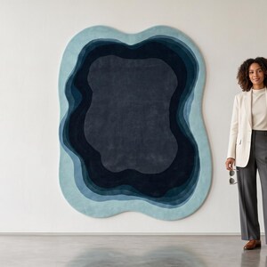 May include: An irregularly shaped wall rug featuring concentric, organic layers in shades of light blue, medium blue, and deep navy. The rug has a soft, textured surface and is mounted on a plain white wall, creating a bold, layered visual effect.