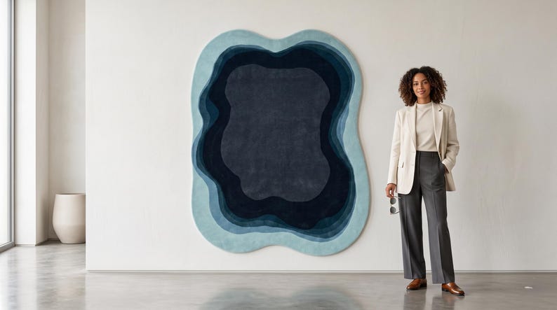 May include: An irregularly shaped wall rug featuring concentric, organic layers in shades of light blue, medium blue, and deep navy. The rug has a soft, textured surface and is mounted on a plain white wall, creating a bold, layered visual effect.
