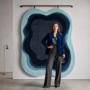 May include: A large, irregular, wall-mounted rug featuring concentric, wavy layers of blue, ranging from light cyan to deep navy. The rug is displayed on a dark horizontal rod against a plain wall, creating a layered, topographical appearance.