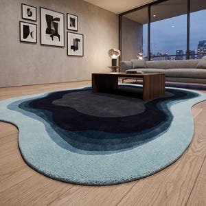 May include: An irregular, organic-shaped area rug featuring concentric, wavy layers of colour. The design transitions from a dark charcoal centre through shades of navy and medium blue, ending in a light, pale blue border on a wooden floor.