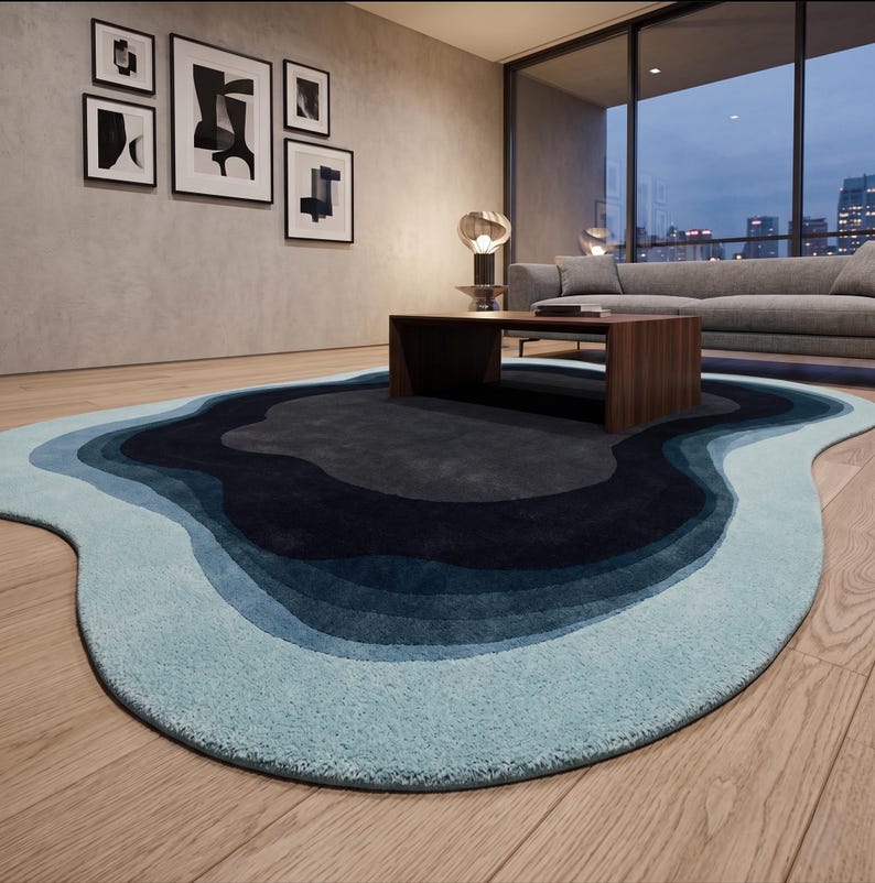 May include: An irregular, organic-shaped area rug featuring concentric, wavy layers of colour. The design transitions from a dark charcoal centre through shades of navy and medium blue, ending in a light, pale blue border on a wooden floor.