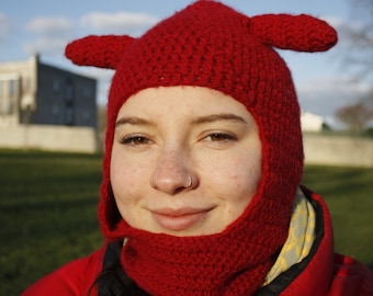 May include: A red, handmade crochet hat with ear-like protrusions and a face covering. The hat is part of a set, and the person in the image is wearing a red jacket. The background includes a blue sky and a building.