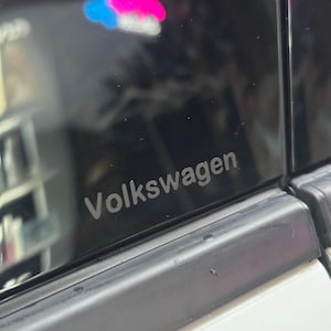 May include: A close-up of a black car window with the word "Volkswagen" printed in silver lettering.