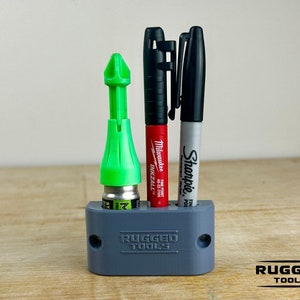 May include: A gray 3D printed holder with the words "RUGGED TOOLS" printed on the front. The holder has three slots, one for a green glue applicator, one for a red Milwaukee marker, and one for a black Sharpie marker.
