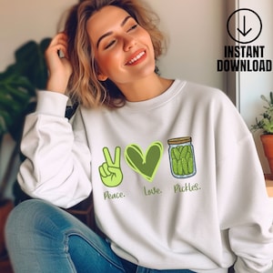Pode incluir: Camisola branca com um sinal de paz verde, um coração verde e um frasco de picles verdes. O texto diz "Peace. Love. Pickles."