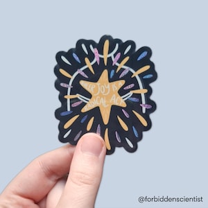 Könnte beinhalten: Ein Aufkleber mit einem schwarzen Hintergrund und einem Sternmuster. Der Aufkleber hat eine zentrale Sternform mit den Worten "Queer Joy is a Radical Act" in einem kreisförmigen Muster. Das Design enthält orange, gelbe, blaue und rosa Akzente.