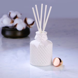 May include: A white, diamond-patterned diffuser bottle with white reeds and a decorative bow. A copper-toned cap sits beside the bottle. A small black bowl with cotton buds is in the background. The items are on a marble surface.