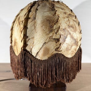 Peut inclure: Lampe de table vintage avec un abat-jour rond marbré beige et marron. L'abat-jour est cousu avec du fil marron et orné d'une frange marron. La lampe repose sur un socle rectangulaire marron foncé.