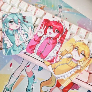 May include: Three anime-style character keycaps with colorful designs. The keycaps feature illustrations of girls in maid outfits, with details like teal, pink, and yellow clothing. The keycaps are on a pink keyboard.