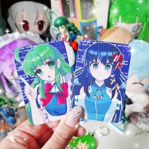 Könnte beinhalten: Zwei Karten mit Anime-Charakteren werden vor einem verschwommenen Hintergrund aus Plüschtieren und Figuren gehalten. Die Karten zeigen weibliche Charaktere mit grünen und blauen Haaren, die blaue und weiße Outfits tragen. Die Karten haben Text in einem blauen Rand.