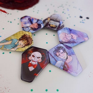 Könnte beinhalten: Sechs sechseckige Pins mit verschiedenen bedruckten Bildern. Die Pins sind in einem Kreis auf einer weißen Oberfläche mit roten und schwarzen Spritzern angeordnet. Die Bilder zeigen einen Mann mit einer Puppe, einen Mann mit einem Verband, einen Mann mit einer blutigen Wunde und einen Mann mit einem Verband.