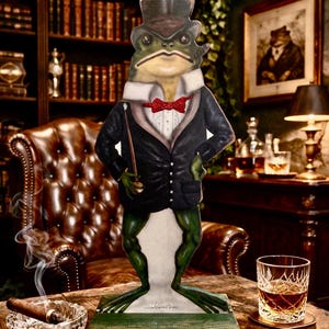May include: A whimsical frog figure dressed in a black suit, top hat, and red bow tie, holding a cane. The frog stands on a green base, with a cigar and a glass of amber liquid nearby. The background includes a leather chair and a bookshelf.