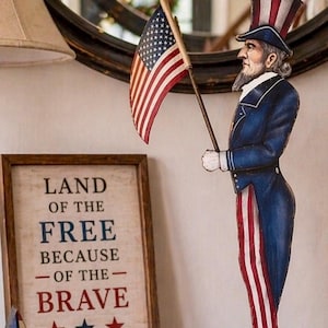 Könnte beinhalten: Eine Holzfigur von Uncle Sam, die eine amerikanische Flagge hält, mit einem Schild mit der Aufschrift „Land of the Free Because of the Brave“. Die Figur trägt einen blauen Mantel, rot-weiß gestreifte Hosen und einen Zylinder. Das Schild hat einen Holzrahmen.