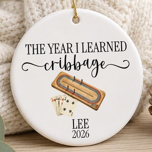 May include: A round white ceramic ornament featuring the black text THE YEAR I LEARNED cribbage, an illustration of a wooden cribbage board with pegs and playing cards, and the black text LEE 2026. A gold string is attached to the top for hanging.