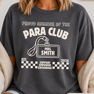 May include: Dark grey t-shirt with the text "PROUD MEMBER OF THE PARA CLUB" and a name tag that says "MRS. SMITH". The shirt also has the text "EST. 2026" and "SUPPORT EMPOWER ENCOURAGE".