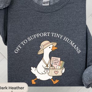 Peut inclure: Un sweat-shirt à col rond gris chiné foncé avec un motif représentant une oie blanche portant un chapeau de soleil beige et un cordon marqué PARA PROFESSIONAL. L'oie porte un panier avec des livres et le texte OFF TO SUPPORT TINY HUMANS est imprimé.