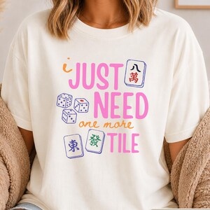 May include: An ivory t-shirt featuring the pink and orange text "I JUST NEED one more TILE" alongside illustrations of three white dice and three mahjong tiles with blue, red, and green symbols. The shirt has a crew neckline and short sleeves.