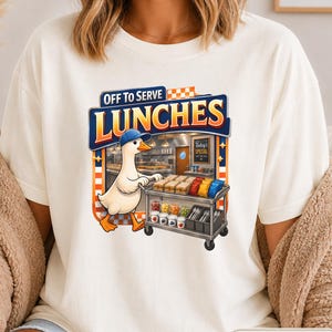 May include: An ivory t-shirt featuring a graphic of a white duck wearing a blue baseball cap, pushing a metal cart filled with sandwiches, snacks, and containers. The text above the duck reads "OFF TO SERVE LUNCHES" in bold blue and orange letters.