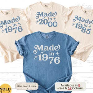 May include: A collection of t-shirts in blue and ivory. Each shirt features the text "Made in" followed by a year, such as 1976, 2000, and 1985, in a retro font. The shirts are available in multiple sizes and colors.