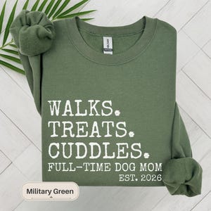 May include: Military green sweatshirt with white text: "WALKS. TREATS. CUDDLES. FULL-TIME DOG MOM EST. 2026." The sweatshirt has a crew neck and long sleeves. The color is labeled "Military Green."