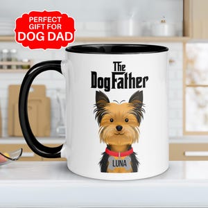 May include: White ceramic mug with a black interior and handle. The mug features the text "The Dogfather" above a cartoon illustration of a Yorkshire Terrier dog with a red collar and a name tag that says "LUNA".