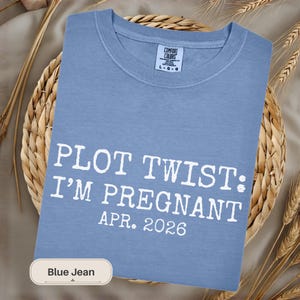 May include: A blue jean Comfort Colors t-shirt with the text "PLOT TWIST: I'M PREGNANT APR. 2026" in white. The shirt is folded on a woven basket with wheat stalks.