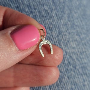 May include: A small, gold-coloured horseshoe charm with a loop for attaching to a necklace or bracelet. The horseshoe has a curved shape with small, evenly spaced indentations along the bottom. The charm is held against a blue denim background.
