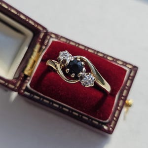 Puede incluir: Un anillo de oro con una piedra oscura en el centro y dos piedras más pequeñas y claras a cada lado. El anillo se muestra en un joyero de terciopelo rojo con detalles dorados.