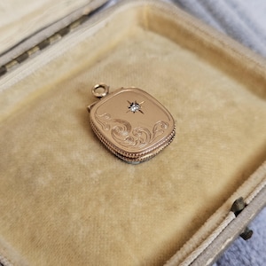 May include: A gold-colored, rectangular locket with a starburst design and a small gemstone in the center. The locket features ornate, swirling engravings and is presented in a beige velvet-lined case.