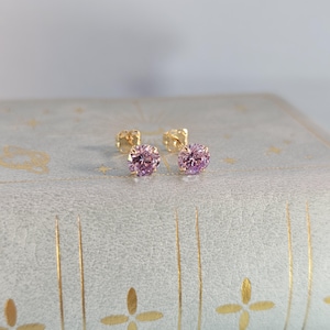 May include: A pair of stud earrings featuring round, light purple gemstones set in gold-colored metal. The earrings are displayed on a light blue surface with gold-colored decorative accents. The earrings are a classic jewelry item.
