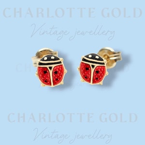May include: A pair of stud earrings shaped like ladybirds with red and black enamel detailing on a gold-tone metal base. The ladybird wings feature a textured grid pattern and black spots. The text CHARLOTTE GOLD Vintage jewellery is visible.