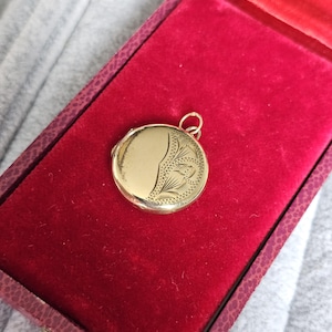 May include: A gold-colored round locket with an etched floral design on a red velvet jewelry box. The locket is open, revealing a space for a photograph or keepsake. The box has a patterned exterior.
