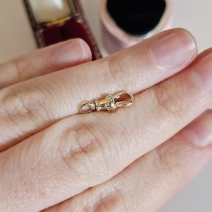 Può includere: Un piccolo fermaglio color oro con un design girevole. Il fermaglio è tenuto tra le dita, con uno sfondo sfocato che include un portagioie e un astuccio foderato di rosso. Il fermaglio è probabilmente per gioielli o un accessorio simile.