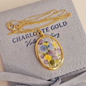 Pode incluir: Um colar dourado com um pingente oval com um design floral em tons de azul, roxo e amarelo. O pingente &eacute; emoldurado em ouro e pendurado em uma delicada corrente de ouro. O colar repousa sobre uma bolsa azul clara com o texto "CHARLOTTE GOLD Vintage Jewelry".