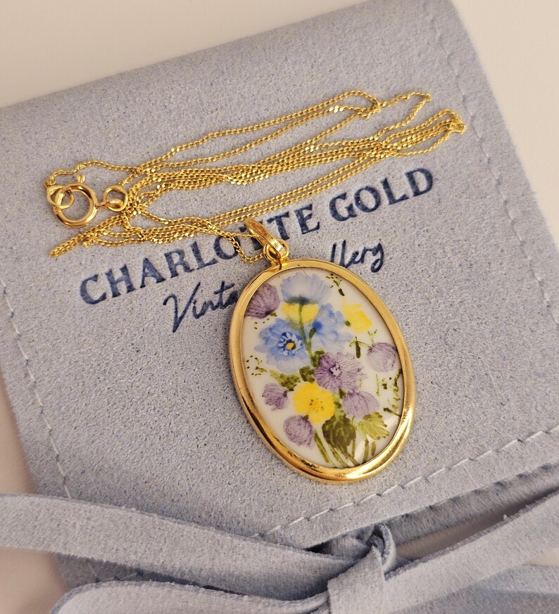 Pode incluir: Um colar dourado com um pingente oval com um design floral em tons de azul, roxo e amarelo. O pingente &eacute; emoldurado em ouro e pendurado em uma delicada corrente de ouro. O colar repousa sobre uma bolsa azul clara com o texto "CHARLOTTE GOLD Vintage Jewelry".