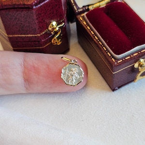 May include: A small, octagonal gold pendant featuring an engraved depiction of Saint Christopher carrying the Christ child. The pendant has a textured, faceted border and a small jump ring at the top for attachment to a chain or necklace.