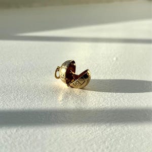 May include: A small, open gold-coloured pendant with a spherical design. The pendant is split in half, revealing a detailed interior. The pendant has a small loop for a chain. The pendant is on a white surface.
