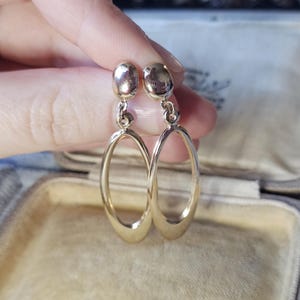May include: A pair of gold oval drop earrings. Each earring features a small, rounded top linked to a larger, elongated oval. The earrings are presented against a soft, neutral background, suggesting a jewellery display.