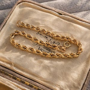 May include: A gold-colored rope chain bracelet displayed in a vintage jewelry box. The bracelet is made of twisted links and has a clasp closure. The jewelry box is beige with a soft, textured interior.
