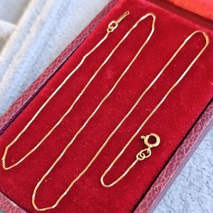 May include: A delicate gold necklace with a box chain design, displayed inside a red velvet jewelry box. The necklace is shown with its clasp open, ready to be worn. The box has a dark red exterior.